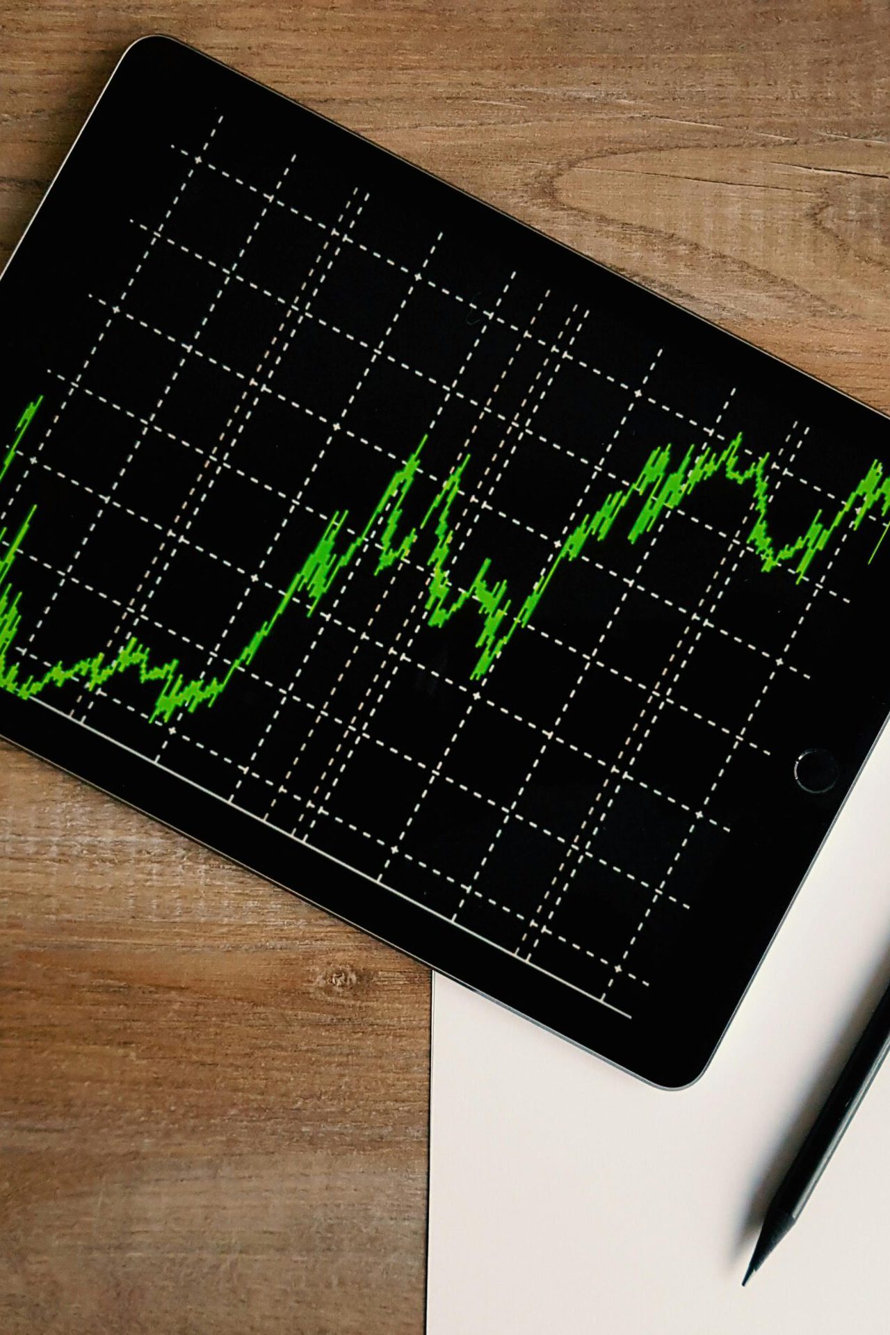 Tablet mit grünem Liniendiagramm auf schwarzem Bildschirm, das steigende Werte zeigt, liegend auf einem Holztisch neben Papier und Stift – Symbol für Datenanalyse und Wachstum.